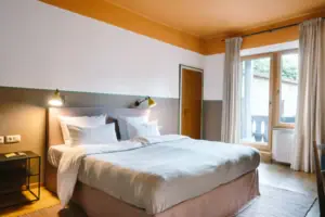 A cosy bed in a modern hotel room at the HENRI Hotel Garmisch-Partenkirchen with access to the balcony in the background.